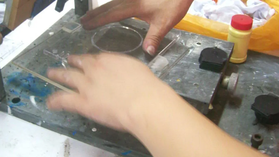 Archived work-instruction photograph showing hands placing a transparent plastic part into a screen-printing locating fixture